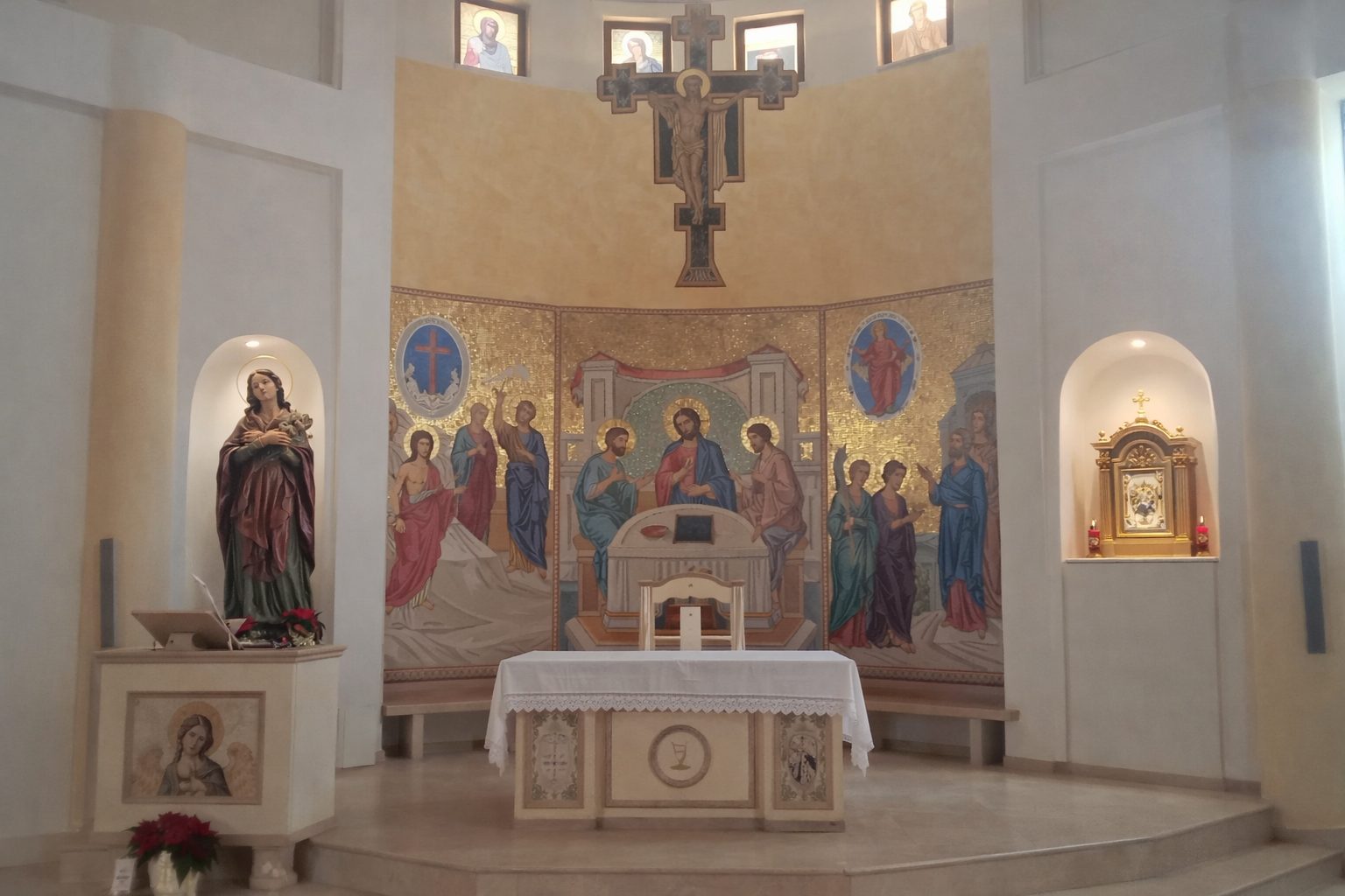 Chiesa Santa Maria Goretti L'abside vista di fronte: l'altare in marmo chiaro davanti al mosaico a fondo oro, il crocifisso ligneo dipinto sopra e le due nicchie con la statua della Madonna e il tabernacolo, nella Chiesa di Santa Maria Goretti