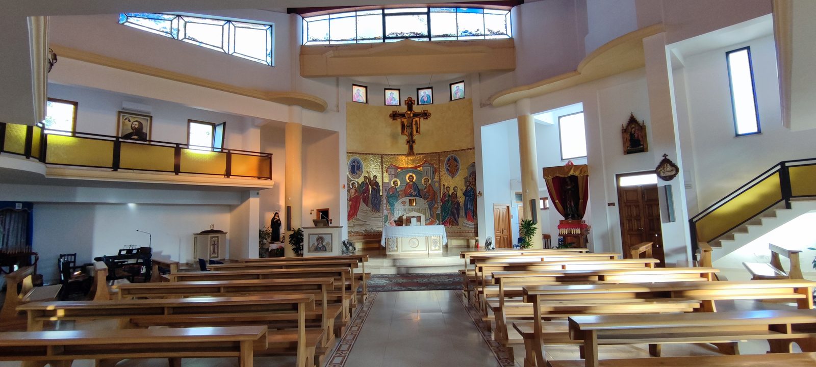Chiesa Santa Maria Goretti L'aula vista dal fondo verso il presbiterio, con i banchi chiari ai lati del corridoio centrale e il mosaico absidale illuminato dalle finestre alte, nella Chiesa di Santa Maria Goretti
