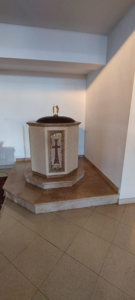 Fonte battesimale ottagonale in pietra chiara, con una croce a mosaico dorato sulla vasca e il coperchio scuro sormontato da una crocetta, nella Chiesa di Santa Maria Goretti