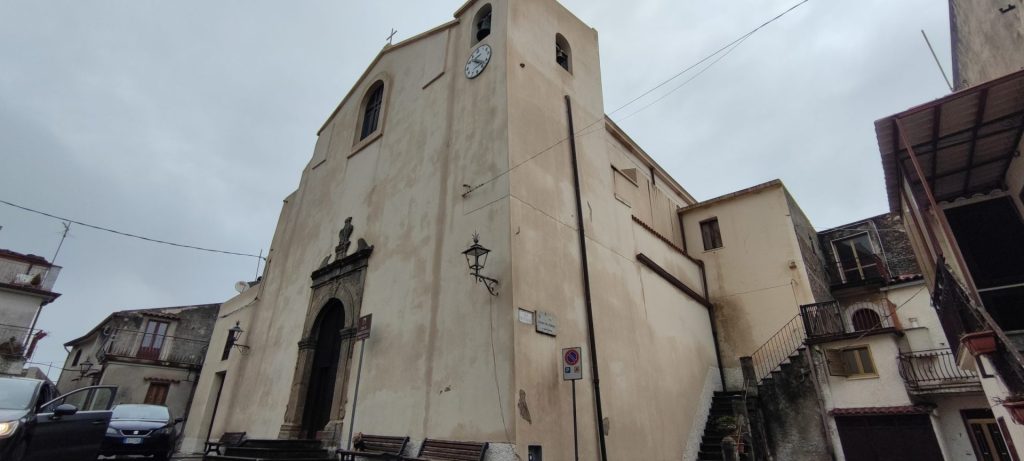 Facciata intonacata chiara della Chiesa Matrice di Sant'Andrea Apostolo, con il portale in pietra, la statua nella nicchia e il campanile con l'orologio, tra le case del centro storico di Guardia Piemontese