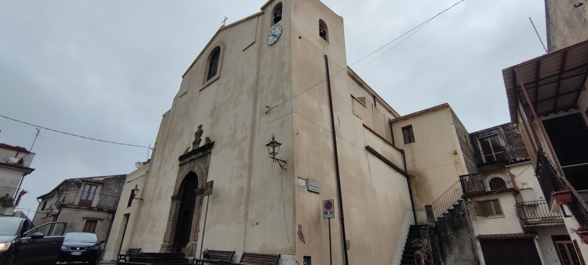 Home Facciata intonacata chiara della Chiesa Matrice di Sant'Andrea Apostolo, con il portale in pietra, la statua nella nicchia e il campanile con l'orologio, tra le case del centro storico di Guardia Piemontese